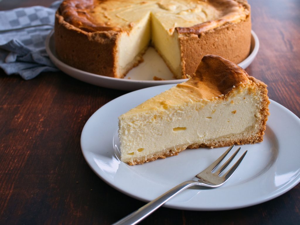 Ein Stück Käsekuchen auf einem Wasser Teller. Im Hintergrund ein ganzer Käsekuchen.