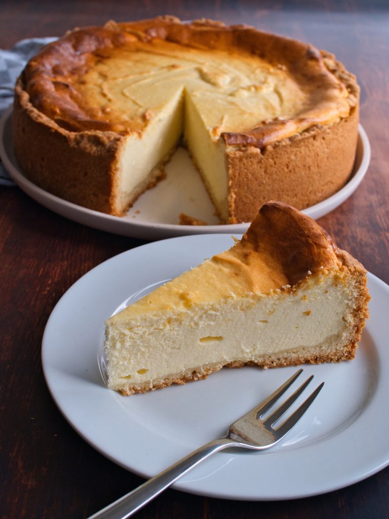 Ein Stück Käsekuchen auf einem Wasser Teller. Im Hintergrund ein ganzer Käsekuchen.