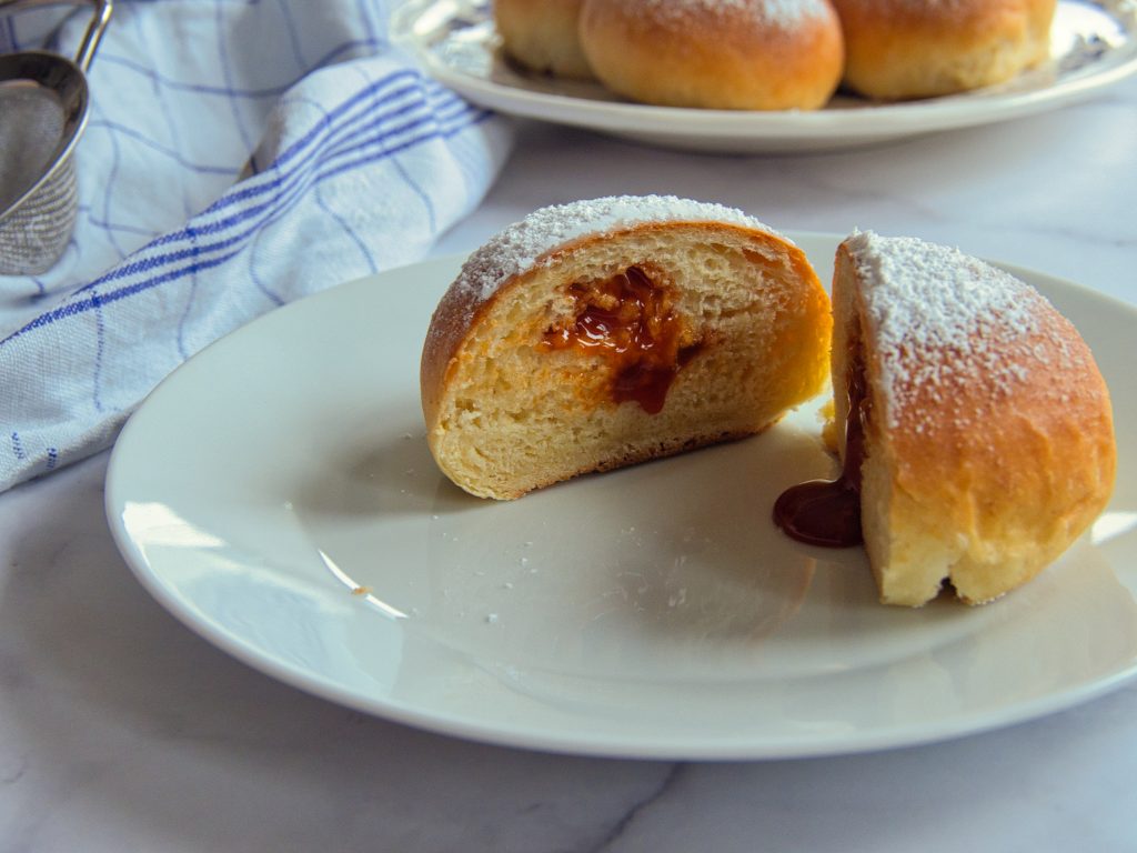 Ein aufgeschnittener Krapfen auf einem Teller bei dem Marmelade herausläuft. Im Hintergrund weitere. Krapfen auf einem Teller.